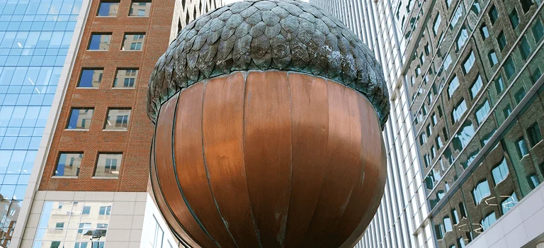 A large bronze acorn sculpture stands between modern glass and brick buildings, reflecting urban art in a cityscape.